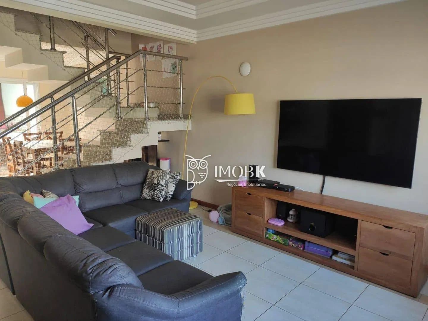 Sala de estar com sofá cinza, rack de madeira, TV e escada ao fundo.