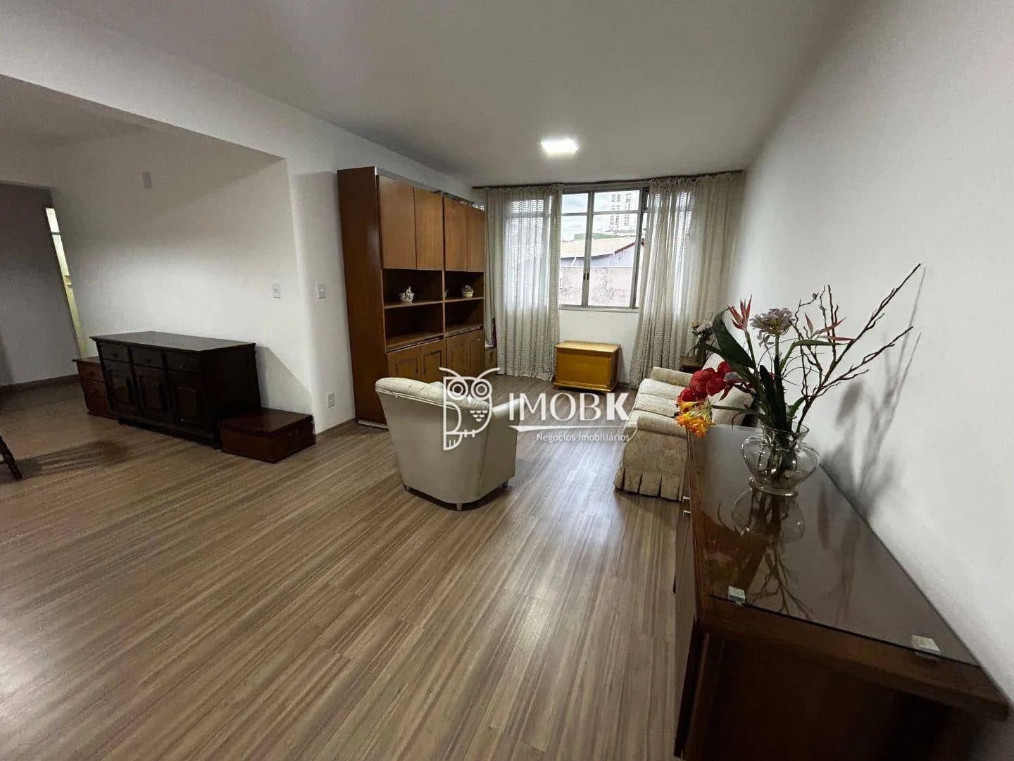 Sala de estar ampla com piso amadeirado, sofá, poltrona e estante de madeira.