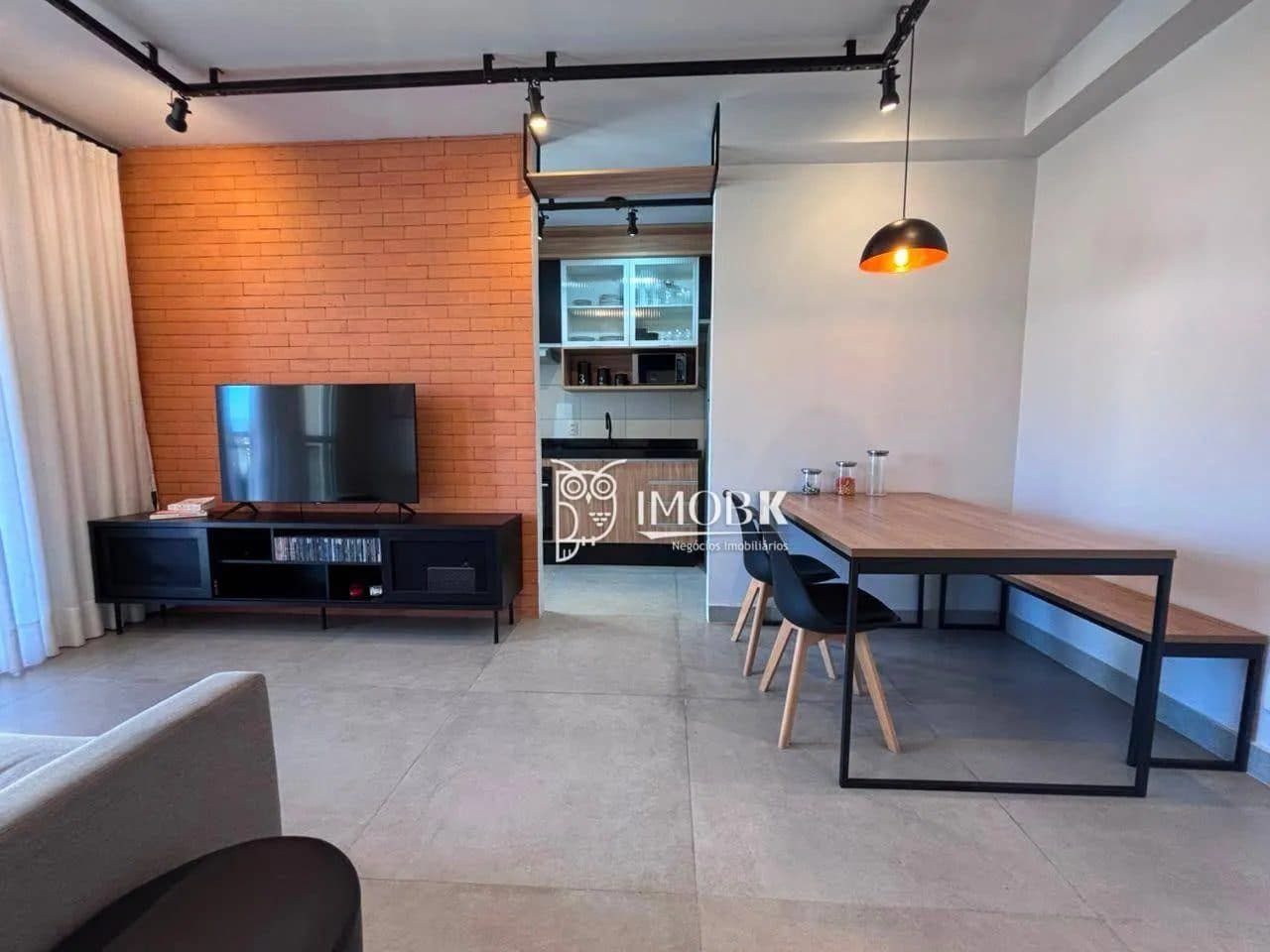 Visão ampla da sala integrada com sofá cinza, rack, mesa de jantar e acesso à varanda com cortinas brancas.