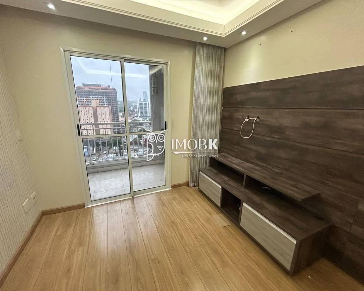 Sala de estar com painel para TV em tom amadeirado, piso laminado e acesso à varanda.