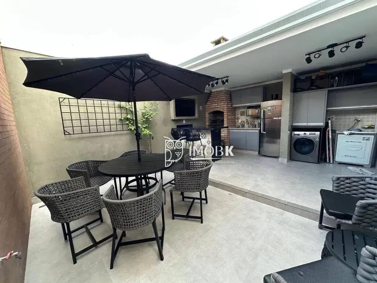 Área gourmet externa com churrasqueira de tijolos à vista, mesa com cadeiras, guarda-sol e pia.